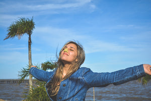 blonde girl in the sun with her eyes closed with arms wide open and wearing colorful reef safe Nöz sunscreen under a blue sky